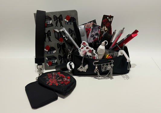 A black fabric organizer pouch set displayed on a white surface, featuring a main bag with a gray bow print, a black zippered pouch with red floral embroidery, and a black card holder with silver chain details. The background shows a white storage organizer filled with colorful pens and stationery.