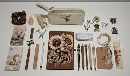 A styled flat-lay display of various stationery and accessory items, featuring a central brown journal with a detailed sunflower and gear illustration, surrounded by pens, a beige zip pouch, a beaded bracelet, and other matching aesthetic items on a white surface.