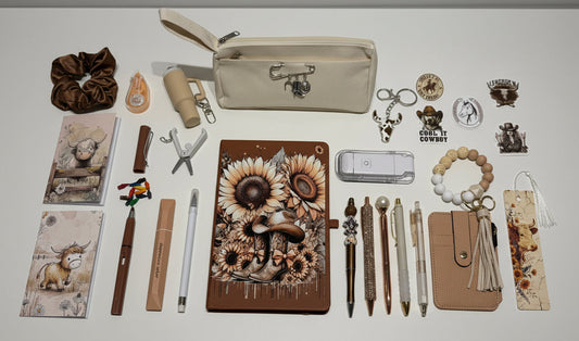 A styled flat-lay display of various stationery and accessory items, featuring a central brown journal with a detailed sunflower and gear illustration, surrounded by pens, a beige zip pouch, a beaded bracelet, and other matching aesthetic items on a white surface.
