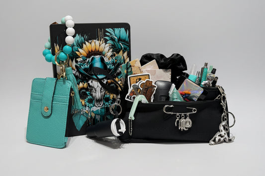 A styled flat-lay of a black canvas organizer pouch filled with stationery and accessories, next to a turquoise leather card holder and a black notebook with a colorful skull graphic, set against a background of white storage drawers filled with colorful pens.