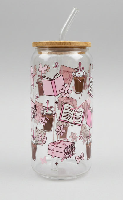  a clear glass tumbler with a bamboo lid, decorated with pink illustrations of books, flowers, and gift boxes, set against a blurred green outdoor background.