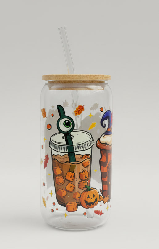 A clear glass tumbler with a bamboo lid and clear straw, decorated with Halloween-themed illustrations including a gummy bear drink, a witch cupcake, pumpkins, and autumn leaves. It is placed on a gray woven patio table with another similar tumbler blurred in the background.