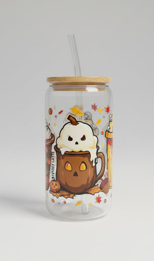 A clear glass can-shaped tumbler with a bamboo lid and clear straw, decorated with a cute cartoon pumpkin mug and ghost design, sitting on a gray woven patio table.