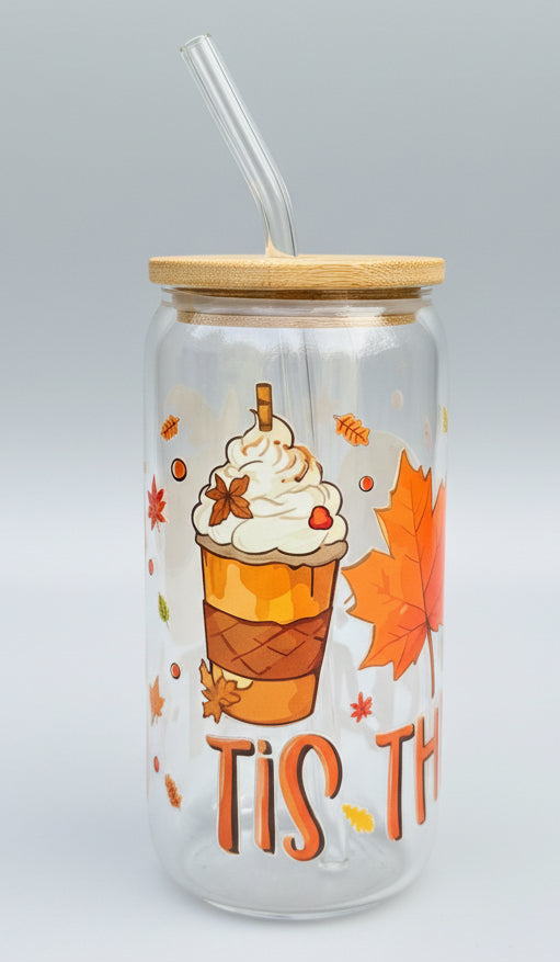A clear glass can-shaped tumbler with a bamboo lid and glass straw, decorated with an autumn theme featuring a fall drink, maple leaves, and the text 'TIS TH'.