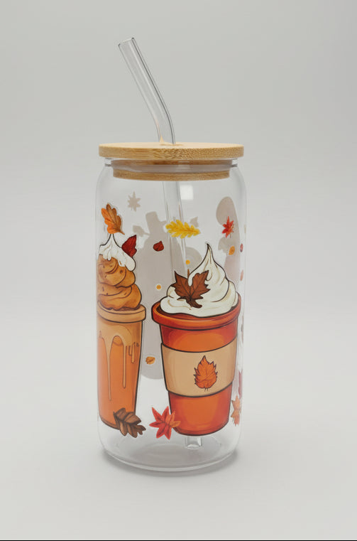 A clear glass can-shaped tumbler with a bamboo lid and clear glass straw, decorated with illustrations of fall drinks, whipped cream, maple leaves, and autumn leaves. The tumbler is placed on a gray woven outdoor patio chair.