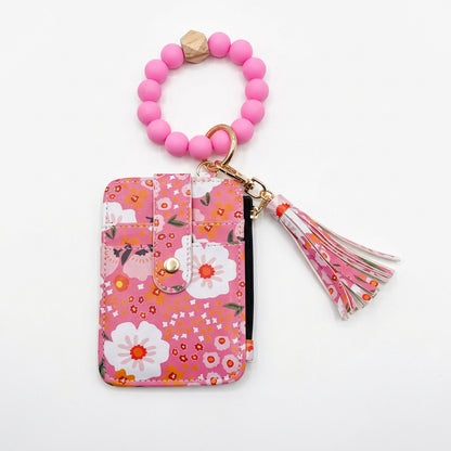 A pink floral card holder wallet with a matching pink beaded wrist strap and tassel charm, displayed on a light gray background.