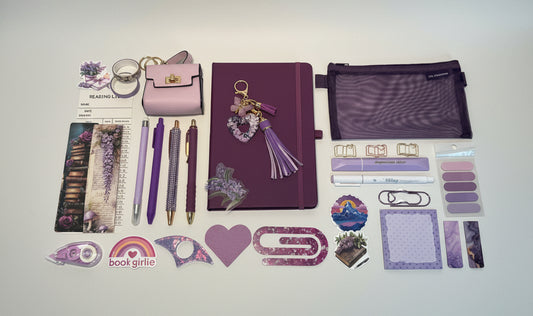 Collection of purple stationery items including pens, a notebook, and decorative elements on a white surface with storage drawers in the background.